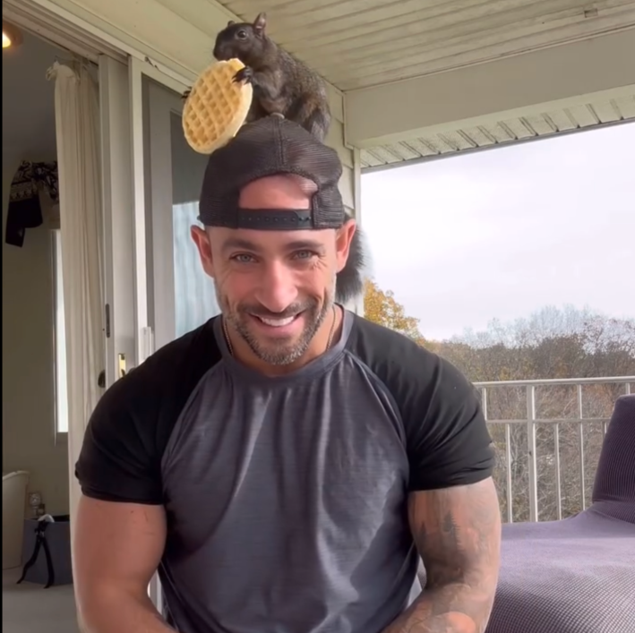 a screenshot of a white man with a backwards baseball cap with a gray squirrel sitting on his head eating a waffle